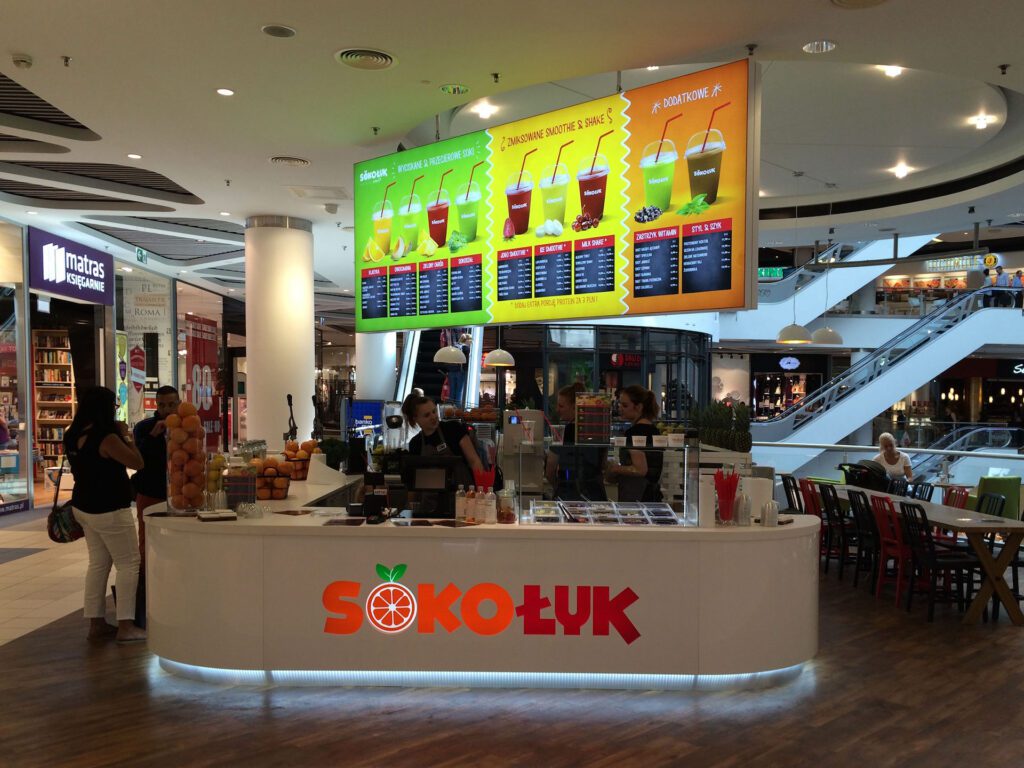 tętniąca życiem wyspa gastronomiczna Redberry-Sokołyk w centrum handlowym, z personelem za ladą, wystawami owoców i kolorowymi tablicami z menu powyżej. W tle widoczni są klienci i część wypoczynkowa. projekt i nadzór Tatjana Kobuszewska trksystem.com