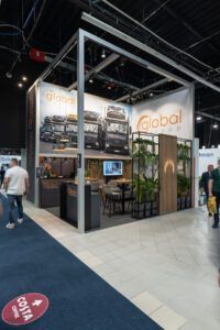 Stoisko targowe Global Motion na Ptak Warsaw Expo prezentuje duże zdjęcia pojazdów, efektowne oznakowanie, recepcję, zielone rośliny i monitor, a po hali wystawowej przechodzą zwiedzający. projekt i nadzór Tatjana Kobuszewska trksystem.com