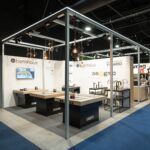 Nowoczesne stoisko targowe 3Sygma Warsaw Home obejmuje drewniane i metalowe ekspozytory kuchenne pod metalową ramą z górnym oświetleniem. Logotypy marek, w tym "benthaus" i "Brand Vario", wyróżniają się na białych ścianach, gdy ludzie wchodzą w interakcje na niebieskim dywanie. projekt i nadzór Tatjana Kobuszewska trksystem.com