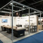 Nowoczesne stoisko targowe 3Sygma na targach Warsaw Home prezentuje zlewozmywaki, baterie i armaturę na ladach. Stoisko ma białe ściany z logo marki, przemysłowe oświetlenie nad głową, niebieski dywan i ludzi widocznych w pobliżu innych stoisk. projekt i nadzór Tatjana Kobuszewska trksystem.com