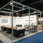 Na stoisku Warsaw Home firmy 3Sygma zlewozmywaki, baterie i armatura kuchenna są prezentowane na eleganckich blatach pod industrialnym oświetleniem. Na białych ścianach widnieją logotypy takich marek jak Benthaus i Armando Vicario. Dwóch mężczyzn w garniturach rozmawia przy przejściu wyłożonym niebieskim dywanem. projekt i nadzór Tatjana Kobuszewska trksystem.com