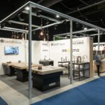 Nowoczesne stoisko targowe na targach 3Sygma Warsaw Home prezentuje armaturę kuchenną i łazienkową, taką jak zlewozmywaki i baterie na drewnianych blatach. Kilka logo marek, w tym Benthaus i Vicario, jest wyświetlanych na białych ścianach, a uczestnicy przechodzą obok po niebieskiej wykładzinie. projekt i nadzór Tatjana Kobuszewska trksystem.com