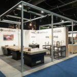 Nowoczesne stoisko wystawiennicze na targach 3Sygma Warsaw Home prezentuje stylowe zlewozmywaki, baterie i blaty kuchenne pod metalową ramą z wiszącymi lampami. Markowe oznakowanie Benthaus, Armando Vicario i innych marek znajduje się na białych ścianach nad niebieską wykładziną podłogową. projekt i nadzór Tatjana Kobuszewska trksystem.com