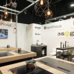 Nowoczesny showroom kuchenny z drewnianymi i czarnymi blatami z wbudowanymi płytami indukcyjnymi, przykładowymi bateriami i geometrycznymi lampami wiszącymi. Na białych ścianach widoczne są znaki firmowe "Benthaus" i "3Sygma Warsaw Home". projekt i nadzór Tatjana Kobuszewska trksystem.com