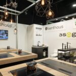 Nowoczesny showroom kuchenny z drewnianymi i czarnymi powierzchniami, płytami indukcyjnymi, garnkami ze stali nierdzewnej, złotymi i czarnymi bateriami, białymi krzesłami, geometrycznymi lampami wiszącymi i logo marek takich jak Benthaus i 3Sygma Warsaw Home na ścianach. projekt i nadzór Tatjana Kobuszewska trksystem.com