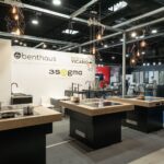 W nowoczesnym showroomie znajdują się wyspy kuchenne z kuchenkami i zlewozmywakami, drewniane blaty oraz wiszące lampy z żarówkami Edisona. Tylną ścianę zdobią szyldy marek benthaus, 3Sygma Warsaw Home i ADAMARO VICARIO. Na wystawie znajduje się kilka baterii. projekt i nadzór Tatjana Kobuszewska trksystem.com