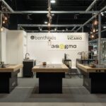 Nowoczesna witryna kuchenna z trzema wyspami wyposażonymi w kuchenki lub zlewozmywaki, pod wiszącymi żarówkami. Branding marek Benthous, Armando Vicario i 3Sygma Warsaw Home widoczny jest na tylnej ścianie. Elegancki, minimalistyczny design. projekt i nadzór Tatjana Kobuszewska trksystem.com