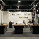 Nowoczesne stoisko targowe 3Sygma Warsaw Home prezentuje trzy minimalistyczne wyspy kuchenne z drewnianymi blatami i czarnymi podstawami. Górne światła zwisają z czarnej ramy, a marki Benthaus i 3Sygma zdobią wyraźne białe ściany. projekt i nadzór Tatjana Kobuszewska trksystem.com