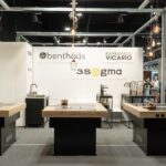 Nowoczesny showroom kuchenny z trzema wyspami kuchennymi, każda z płytą grzewczą i drewnianym blatem, na tle białej ściany z logo "Benthaus", "3Sygma Warsaw Home" i "Armando Vicario". Powyżej wiszą lampy wiszące, a w tle wyświetlane są baterie. projekt i nadzór Tatjana Kobuszewska trksystem.com