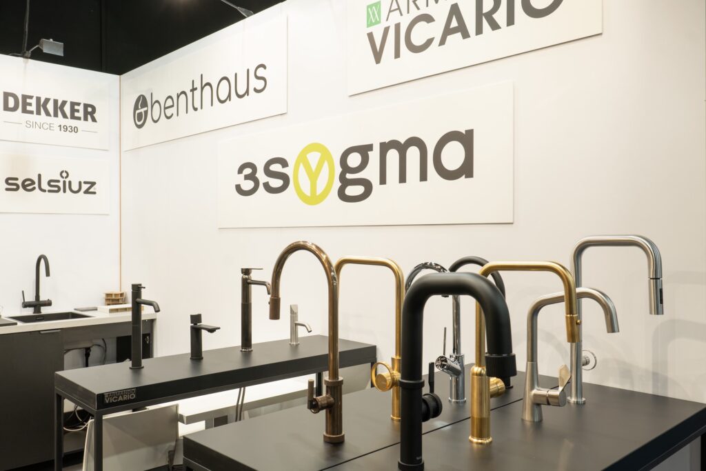 Showroom 3Sygma Warsaw Home prezentuje nowoczesne baterie kuchenne w czarnych, złotych i srebrnych wykończeniach na eleganckich blatach. Kilka znaków firmowych - "benthaus", "3sygma", "DEKKER", "selsius" i "VICARIO" - zdobi białe ściany w tle. projekt i nadzór Tatjana Kobuszewska trksystem.com