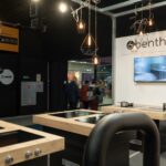 Nowoczesny showroom kuchenny w 3Sygma Warsaw Home posiada drewniane blaty, wbudowane kuchenki i garnki. Powyżej wiszą geometryczne lampy wiszące, a na ekranie ściennym wyświetlane jest logo benthauss. Ludzie i wyświetlacze są widoczne przez szklaną ścianę w tle. projekt i nadzór Tatjana Kobuszewska trksystem.com