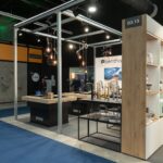 Nowoczesne stoisko targowe z brandingiem SMEG prezentuje urządzenia kuchenne na otwartych półkach na targach 3Sygma Warsaw Home. Centralne lady pokazują baterie i zlewozmywaki pod wiszącymi lampami. W tle przechadzają się ludzie, dodając zorganizowanej, współczesnej atmosfery. projekt i nadzór Tatjana Kobuszewska trksystem.com