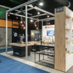 Nowoczesne stoisko targowe na targach 3Sygma Warsaw Home obejmuje urządzenia kuchenne SMEG na białych półkach, drewniane blaty z bateriami i lampy wiszące nad nimi. W tle przechadzają się ludzie na niebieskim dywanie, a wokół znajdują się znaki i banery. projekt i nadzór Tatjana Kobuszewska trksystem.com