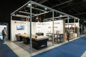 Nowoczesne stoisko targowe z wystawionymi zlewozmywakami, bateriami i armaturą kuchenną. Na białych ścianach widnieją logotypy marek BENTHAUS i VICARIO. Nad nimi wiszą lampy wiszące. projekt i nadzór Tatjana Kobuszewska. Odwiedzający przechodzą po niebieskiej wykładzinie podłogowej. projekt i nadzór Tatjana Kobuszewska trksystem.com