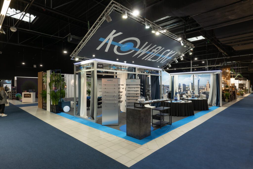 Nowoczesne narożne stoisko wystawowe z nazwą „Kowalski” na dużym, kątowym szyldzie nad głową. Zbudowane jako stoisko targowe z Systemu Octanorm, posiada ekspozycje, stoły i stoiska na niebiesko-białej podłodze w przestronnej, dobrze oświetlonej hali wystawowej. Targi Warsaw Home. projekt i nadzór Tatjana Kobuszewska trksystem.com