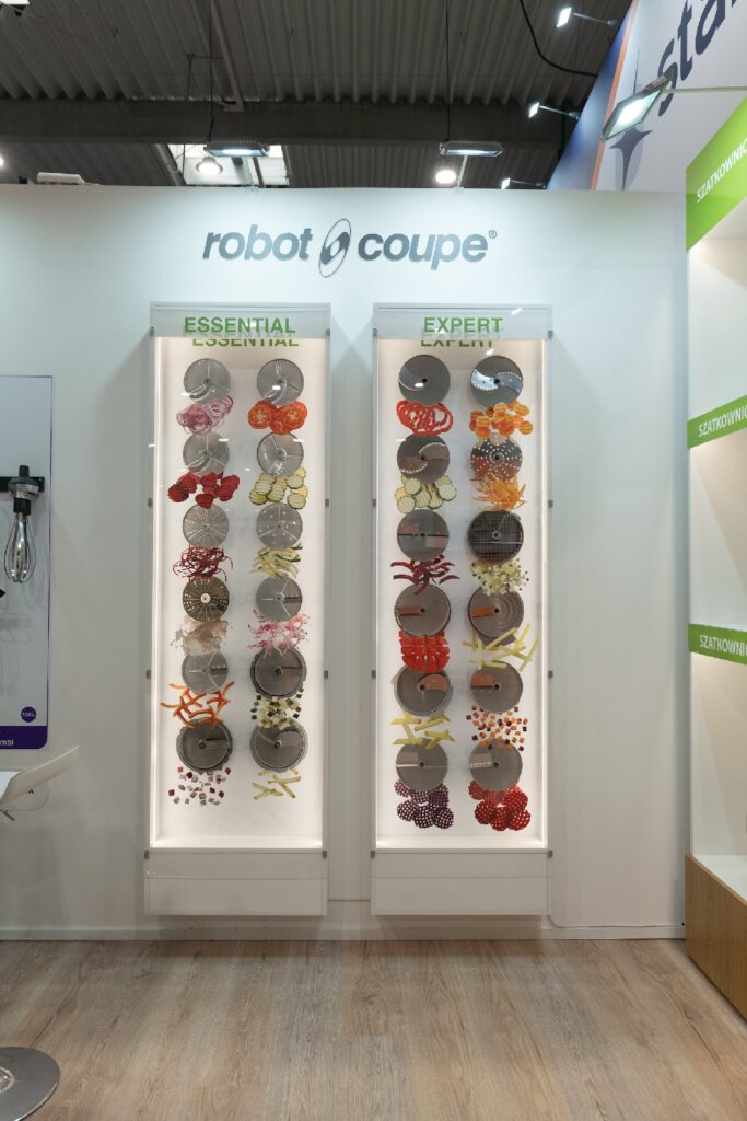 Na dwóch białych panelach wystawowych widoczne są okrągłe ostrza do robotów kuchennych z napisami „ESSENTIAL” i „EXPERT”, sygnowane marką Robot Coupe na targach EuroGastro. Na każdym panelu widnieje wiele błyszczących metalowych ostrzy o różnych kształtach i wycięciach, ułożonych w pionowe rzędy na podświetlonym tle. projekt i nadzór Tatjana Kobuszewska