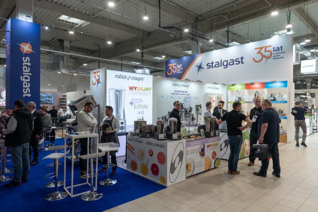 Ludzie rozmawiają przy zatłoczonym stoisku targowym, na którym prezentowany jest sprzęt kuchenny. Widoczne są logo firm Stalgast i Robot Coupe, co podkreśla obecność Robot Coupe na targach EuroGastro. Na białych blatach stoją blendery i rozdrabniacze, a wokół tego obszaru rozmieszczone są banery informacyjne i plakaty produktowe. Scenę oświetlają lampy sufitowe. projekt i nadzór Tatjana Kobuszewska