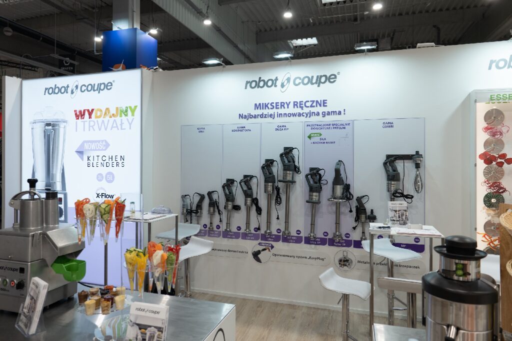 Na dobrze oświetlonym stoisku targowym na białej ścianie wyeksponowano kilka profesjonalnych blenderów ręcznych z napisem „Robot Coupe”. Stoisko, na którym prezentowane są najnowsze innowacje firmy Robot Coupe podczas targów EuroGastro, zawiera również miksery, blendery, roboty kuchenne, kolorowe przybory kuchenne oraz plakaty promocyjne w języku polskim. projekt i nadzór Tatjana Kobuszewska