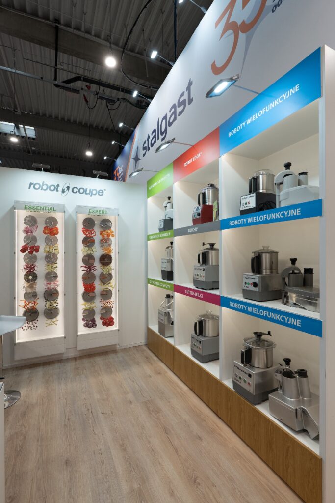 Nowoczesny salon z urządzeniami kuchennymi, w którym na kolorowych półkach wystawiono roboty kuchenne marki Robot Coupe; na lewej ścianie wiszą plakaty przedstawiające składniki, podłoga jest drewniana, a nad głowami wiszą lampy sufitowe, a nad nimi widnieje biało-niebieski szyld Stalgast — wszystko to doskonale oddaje elegancki klimat stoiska Robot Coupe na targach EuroGastro. projekt i nadzór Tatjana Kobuszewska