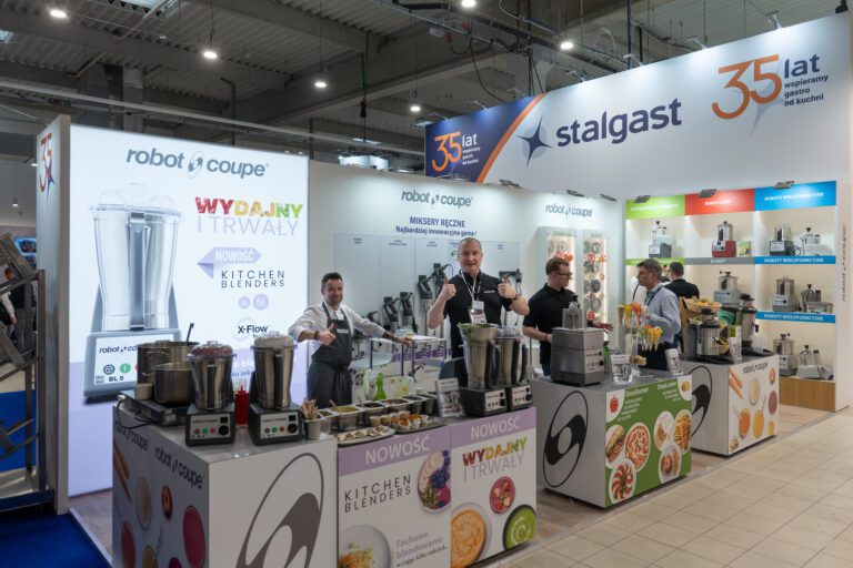 Cztery osoby, w tym Tatjana Kobuszewska, stoją za stoiskiem prezentującym urządzenia kuchenne na targach. Kolorowe banery i nowoczesne wyświetlacze podkreślają blendery i roboty kuchenne, a projekt i nadzór trksystem.com widnieje na oznakowaniu. projekt i nadzór Tatjana Kobuszewska trksystem.com