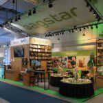 Stoisko targowe firmy Bagstar, zaprojektowane w ramach projektu stoiska i nadzór trksystem.com przez Tatjanę Kobuszewską, wyposażone jest w półki z ekologicznymi opakowaniami, ekran telewizyjny, próbki produktów na stołach i duże szyldy reklamowe pod jasnym oświetleniem w hali wystawowej. projekt i nadzór Tatjana Kobuszewska trksystem.com