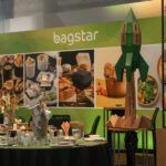 Stoisko targowe Bagstar autorstwa Tatjany Kobuszewskiej zawiera zdjęcia żywności na zielonym tle, ekspozytor w kształcie rakiety oraz okrągły stół z talerzami i pojemnikami. Ciepłe oświetlenie podkreśla ekologiczne opakowania. projekt i nadzór Tatjana Kobuszewska trksystem.com