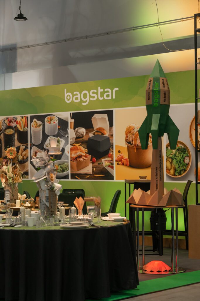 Stoisko targowe Bagstar autorstwa Tatjany Kobuszewskiej zawiera zdjęcia żywności na zielonym tle, ekspozytor w kształcie rakiety oraz okrągły stół z talerzami i pojemnikami. Ciepłe oświetlenie podkreśla ekologiczne opakowania. projekt i nadzór Tatjana Kobuszewska trksystem.com
