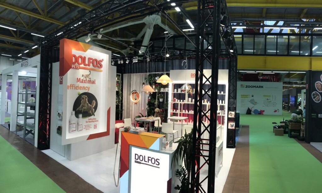 Stoisko targowe DOLFOS PETS z ekspozycjami produktów, plakatami i recepcją. Zaprojektowane przez Tatjanę Kobuszewską z projekt stoiska i nadzór trksystem.com. Jasne oświetlenie, kolorowe dekoracje i sąsiadujące stoiska wypełniają przestronną halę wystawową. projekt i nadzór Tatjana Kobuszewska trksystem.com