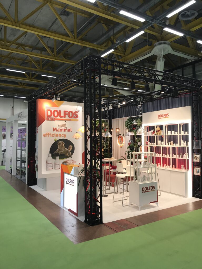 Stoisko targowe narożne firmy Dolfos Pets prezentuje produkty dla zwierząt na półkach z napisem "Maksymalna wydajność" oraz wizerunkami kota i psa. Na stoisku znajdują się krzesła, stoły, oświetlenie i rośliny ozdobne. projekt i nadzór Tatjana Kobuszewska trksystem.com