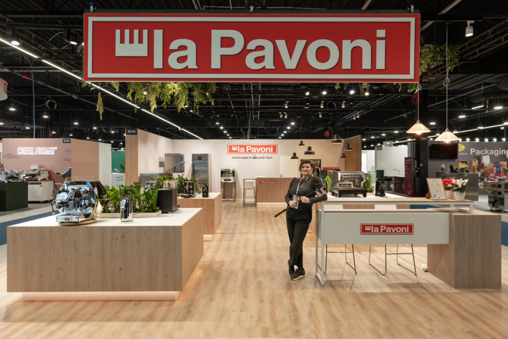 Tatjana Kobuszewska stoi przed stoiskiem targowym La Pavoni firmy Smeg z ekspresami do kawy, roślinami i urządzeniami kuchennymi na drewnianych ladach - świetny przykład dobrze dobranego oświetlenia. projekt i nadzór Tatjana Kobuszewska trksystem.com