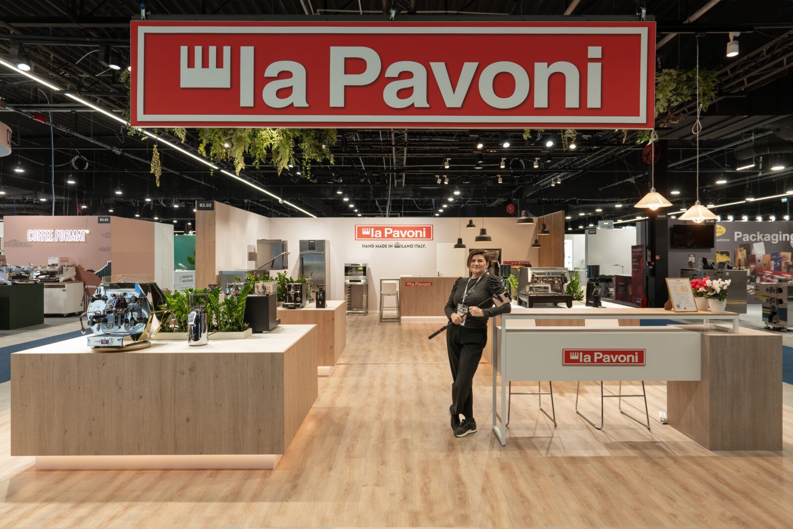 Tatjana Kobuszewska stoi przed stoiskiem targowym La Pavoni firmy Smeg z ekspresami do kawy, roślinami i urządzeniami kuchennymi na drewnianych ladach - świetny przykład dobrze dobranego oświetlenia. projekt i nadzór Tatjana Kobuszewska trksystem.com