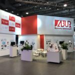 Nowoczesne narożne stoisko wystawiennicze ABUR Systemy Automatyki, zaprojektowane przez projekt stoiska i nadzór trksystem.com Tatjana Kobuszewska, charakteryzuje się białymi ścianami z czerwonymi akcentami, dużym logo, cyfrowymi wyświetlaczami, broszurami, roślinami i ekspozycją produktów automatyki. projekt i nadzór Tatjana Kobuszewska trksystem.com