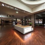 Nowoczesne wnętrze centrum handlowego z drewnianymi podłogami i zakrzywionymi balustradami. W centrum znajduje się biały kiosk ze znakiem Stolz Home, roślinami i cyfrowym ekranem. Sklepy znajdują się w tle, a obszar jest jasno oświetlony naturalnym i sufitowym światłem. projekt i nadzór Tatjana Kobuszewska trksystem.com