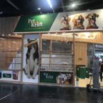 Stoisko targowe Pet Kraft Natural Dog Treats na Interzoo Norymberga zawiera duże zdjęcia szczęśliwych psów, naturalnej wielkości wizerunek psa trzymającego smakołyk, zielony i drewniany wystrój oraz zachęcające ekspozycje produktów w hali wystawowej. projekt i nadzór Tatjana Kobuszewska trksystem.com