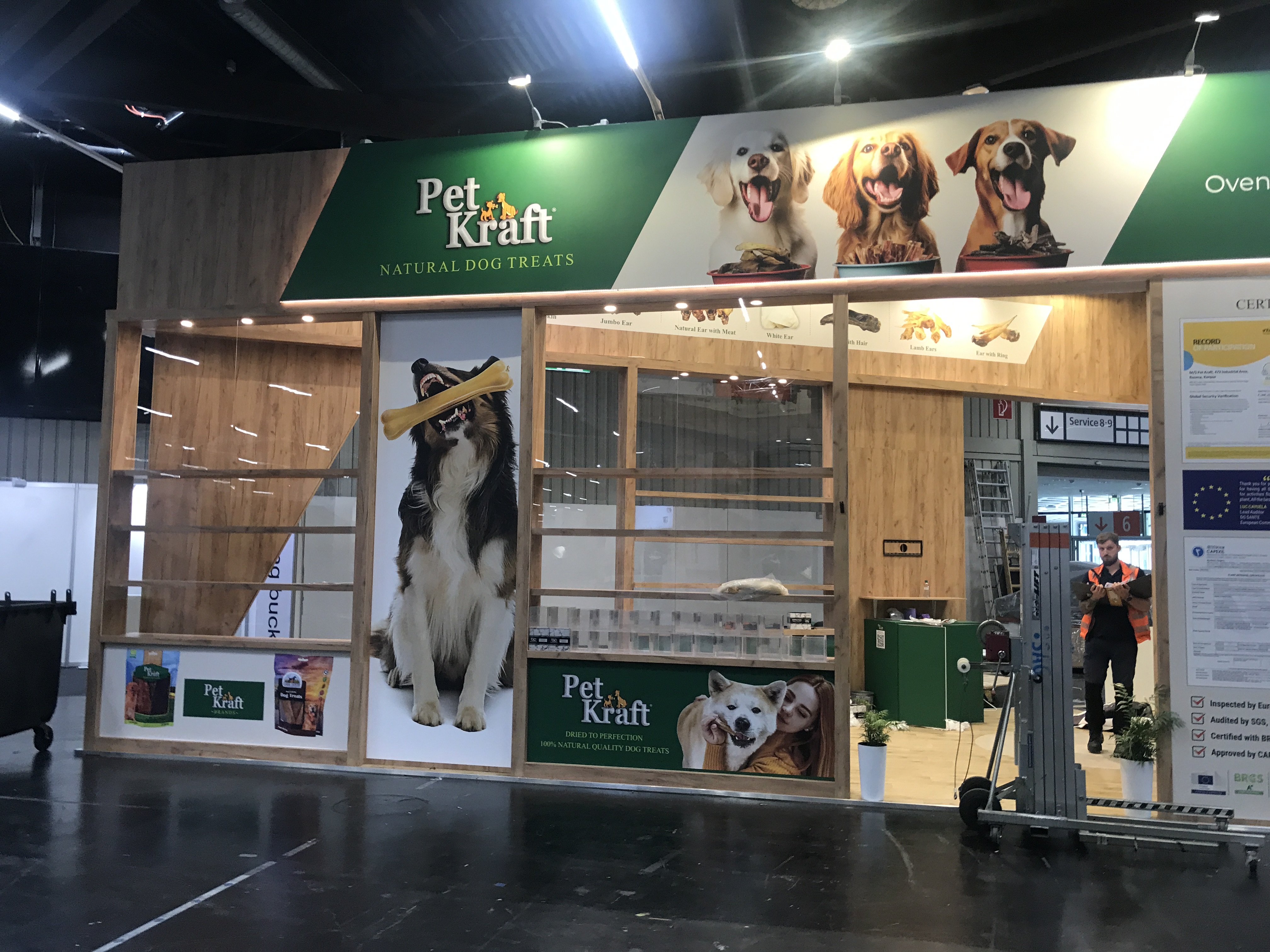 Stoisko targowe Pet Kraft Natural Dog Treats na Interzoo Norymberga zawiera duże zdjęcia szczęśliwych psów, naturalnej wielkości wizerunek psa trzymającego smakołyk, zielony i drewniany wystrój oraz zachęcające ekspozycje produktów w hali wystawowej. projekt i nadzór Tatjana Kobuszewska trksystem.com