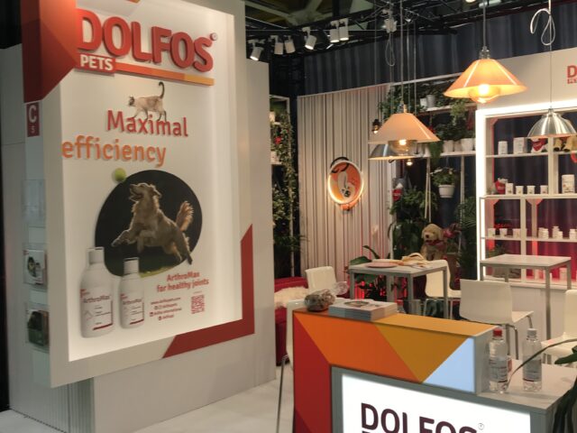 Stoisko targowe firmy DOLFOS PETS zawiera jasne oznakowanie z hasłem "Maksymalna wydajność". Stoisko prezentuje produkty uzupełniające dla zwierząt domowych, podświetlane półki, wiszące lampy i wizerunki psów, a wszystko to w odcieniach bieli, pomarańczy i czerwieni. projekt i nadzór Tatjana Kobuszewska trksystem.com
