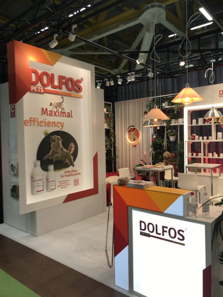 Stoisko targowe firmy DOLFOS PETS zawiera jasne oznakowanie z hasłem "Maksymalna wydajność". Stoisko prezentuje produkty uzupełniające dla zwierząt domowych, podświetlane półki, wiszące lampy i wizerunki psów, a wszystko to w odcieniach bieli, pomarańczy i czerwieni. projekt i nadzór Tatjana Kobuszewska trksystem.com