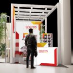 Mężczyzna w czarnej kurtce stoi przed nowoczesnym stoiskiem wystawowym z napisem "DOLFOS" z białymi, czerwonymi i żółtymi akcentami. Na stoisku znajdują się półki z produktami i wysoka szklana konstrukcja przedstawiająca różne zielone rośliny po lewej stronie. projekt i nadzór Tatjana Kobuszewska trksystem.com
