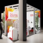 Mężczyzna z teczką stoi w pobliżu nowoczesnego, otwartego stoiska wystawowego Dolfos Pets. Stoisko ma czerwony, żółty i biały branding, półki z produktami, recepcję, zieleń i duży filar częściowo zasłaniający widok. projekt i nadzór Tatjana Kobuszewska trksystem.com