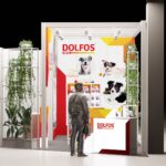 Stoisko targowe firmy DOLFOS charakteryzuje się żywym czerwono-żółtym brandingiem, dużymi zdjęciami psów na tylnej ścianie oraz ladą z ekspozycją produktów. Przy wejściu stoją dwa srebrne posągi ludzi. Zielone rośliny otaczają obie strony stoiska. projekt i nadzór Tatjana Kobuszewska trksystem.com