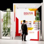 Osoba stoi przed nowoczesnym stoiskiem wystawowym firmy DOLFOS, z białymi ścianami z czerwonymi i żółtymi akcentami, półkami z butelkami i wysokimi zielonymi roślinami do dekoracji. Stoisko jest jasno oświetlone i ma czysty, profesjonalny wygląd. projekt i nadzór Tatjana Kobuszewska trksystem.com