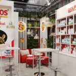 Nowoczesne stoisko wystawowe z czerwono-białym wystrojem obejmuje półki z produktami dla zwierząt domowych, wysokie stoły z przezroczystymi stołkami i przytulne czerwone krzesła. Zielone rośliny i zabawne grafiki zwierząt zdobią przestrzeń, a lampy wiszące zwisają z sufitu. projekt i nadzór Tatjana Kobuszewska trksystem.com