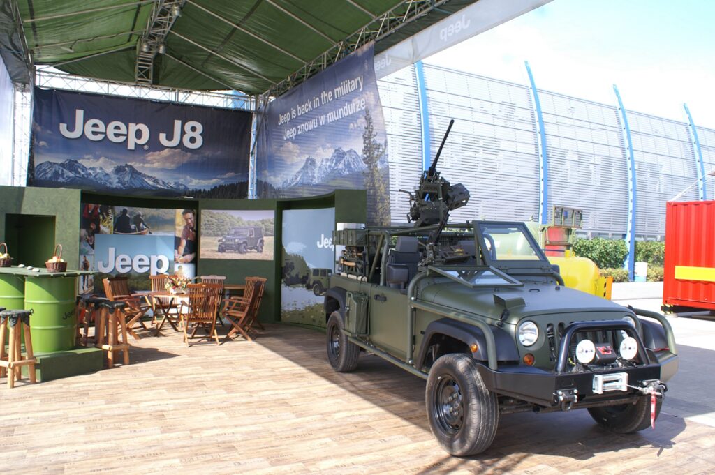 Zielony Jeep J8 w wojskowym stylu z zamontowanym wyposażeniem jest prezentowany pod namiotem z drewnianą podłogą. Za Jeepem znajdują się banery promocyjne Jeepa, meble ogrodowe i drewniana beczka. Tło obejmuje zakrzywione metalowe ogrodzenie i czerwony kontener transportowy. projekt i nadzór Tatjana Kobuszewska trksystem.com