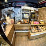 W Przystanku Piekarnia drewniane lady za szkłem eksponują różnorodne pieczywo i wypieki. Bochenki, bułki i rogale opatrzone są etykietami. Widoczne są ceglane i wzorzyste ściany, czarne oznakowanie, krajalnica do chleba, kasa fiskalna i piekarnik. projekt i nadzór Tatjana Kobuszewska trksystem.com