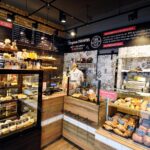 Przytulna piekarnia Przystanek Piekarnia z drewnianym i czarnym wystrojem posiada przeszklone witryny z ciastami i pieczywem. Nad nimi wisi kredowe menu z polskim tekstem, jak widać na fotorelacji Tatjany Kobuszewskiej. Ciepłe oświetlenie tworzy przyjazną atmosferę. projekt i nadzór Tatjana Kobuszewska trksystem.com