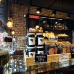 Przytulne wnętrze kawiarni Przystanek Piekarnia z ceglanymi ścianami i przeszkloną witryną, na której znajdują się słoiki z konfiturami, ziołami i wypiekami. Nad ladą wisi kredowe menu, a zielona roślina doniczkowa dodaje uroku temu fotoreportażowi autorstwa Tatjany Kobuszewskiej. projekt i nadzór Tatjana Kobuszewska trksystem.com