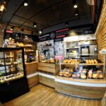 Przytulna piekarnia Przystanek Piekarnia z odsłoniętymi ceglanymi ścianami i ciepłym oświetleniem. Ciasta i pieczywo są wyeksponowane w szklanych gablotach na drewnianych ladach, tworząc rustykalny klimat idealny do fotorelacji autorstwa Tatjany Kobuszewskiej. projekt i nadzór Tatjana Kobuszewska trksystem.com