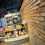 Przy ceglanej ścianie stoi witryna piekarni wypełniona chlebem, rogalikami i ciastkami. Za ladą Przystanku Piekarnia znajdują się piece, ekspresy do kawy i tablica z menu. Sklep emanuje przytulną, nowoczesną atmosferą - idealną na fotorelację autorstwa Tatjany Kobuszewskiej. projekt i nadzór Tatjana Kobuszewska trksystem.com