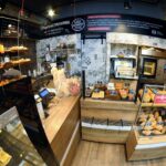 Niewielka, nowoczesna piekarnia Przystanek Piekarnia posiada drewniane i szklane witryny wypełnione pieczywem i wypiekami. Ceglane ściany i wiszące światła tworzą przytulny klimat, gdy pracownik stoi za ladą - idealny do fotorelacji autorstwa Tatjany Kobuszewskiej. projekt i nadzór Tatjana Kobuszewska trksystem.com