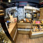 Przytulna piekarnia Przystanek Piekarnia z ceglanymi ścianami i drewnianymi półkami wystawia różne chleby i ciasta za szklanymi ladami. Piece i składniki są widoczne, a na ścianach widnieją polskie znaki. Atmosfera jest ciepła - idealna na fotorelację autorstwa Tatjany Kobuszewskiej. projekt i nadzór Tatjana Kobuszewska trksystem.com